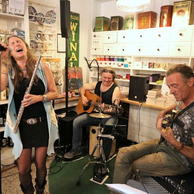 Eine Sängerin mit Querflöte in der Hand und zwei Gitarristen geben in einem Ladenlokal ein Konzert.