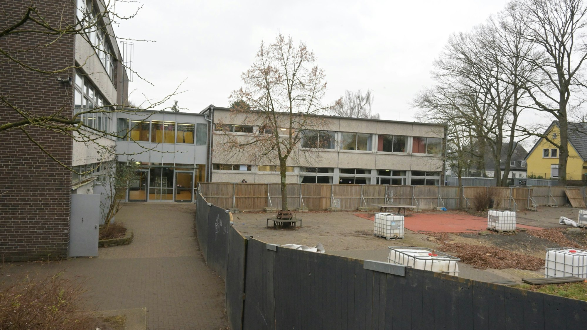 Zwischen dem Schulgebäude und dem Bauzaun sieht man den Gang. Hinter dem Zaun sieht man, den früheren Schulhof.