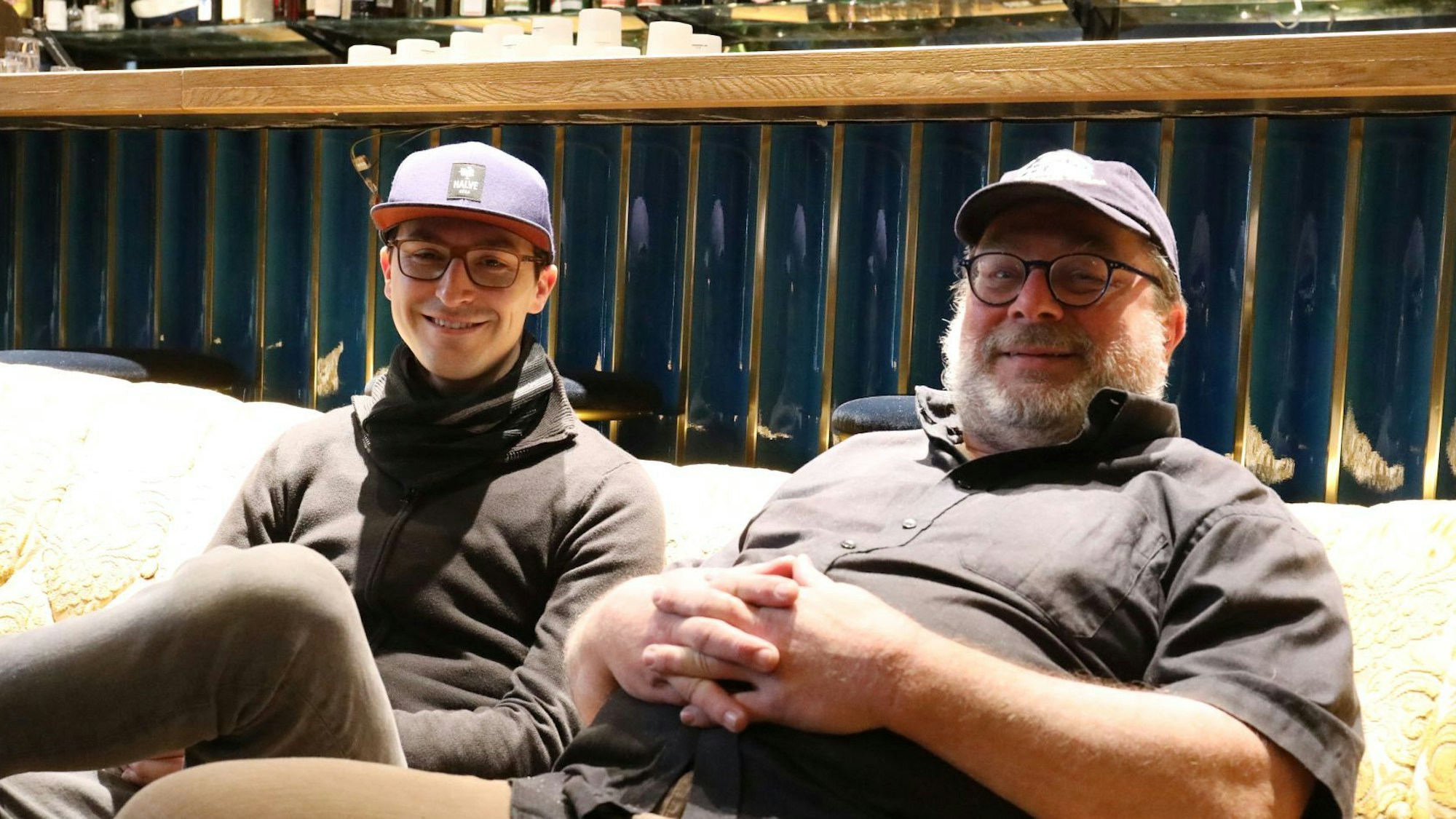 Zwei Männer mit Basecaps sitzen auf einem Sofa vor einer Theke.