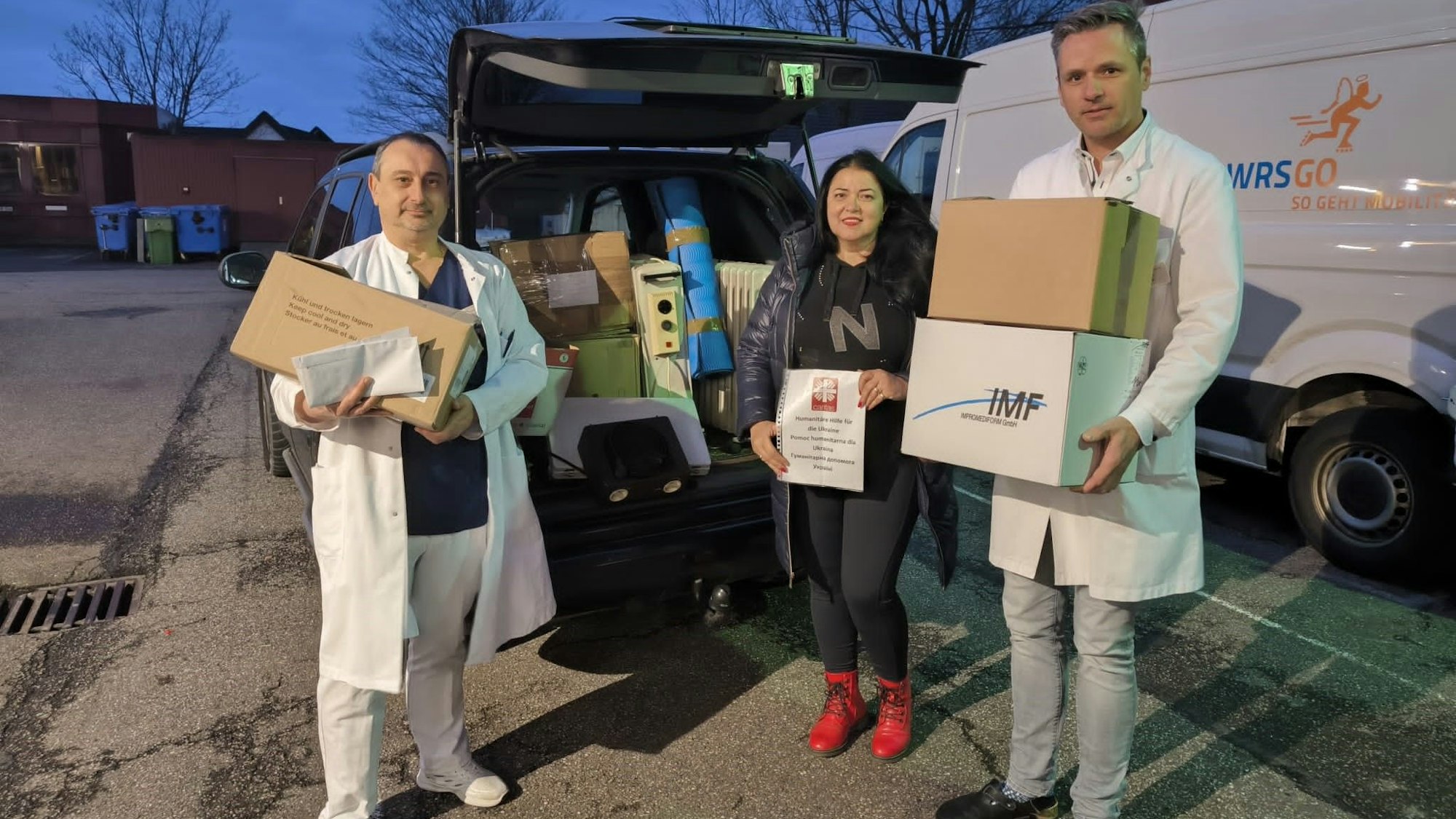 Zwei Männer und eine Frau stehen vor einem Krankenhaus. Hinter ihnen ist ein Geländewagen zu sehen. Sie halten Kartons mit Hilfsmitteln in den Händen.