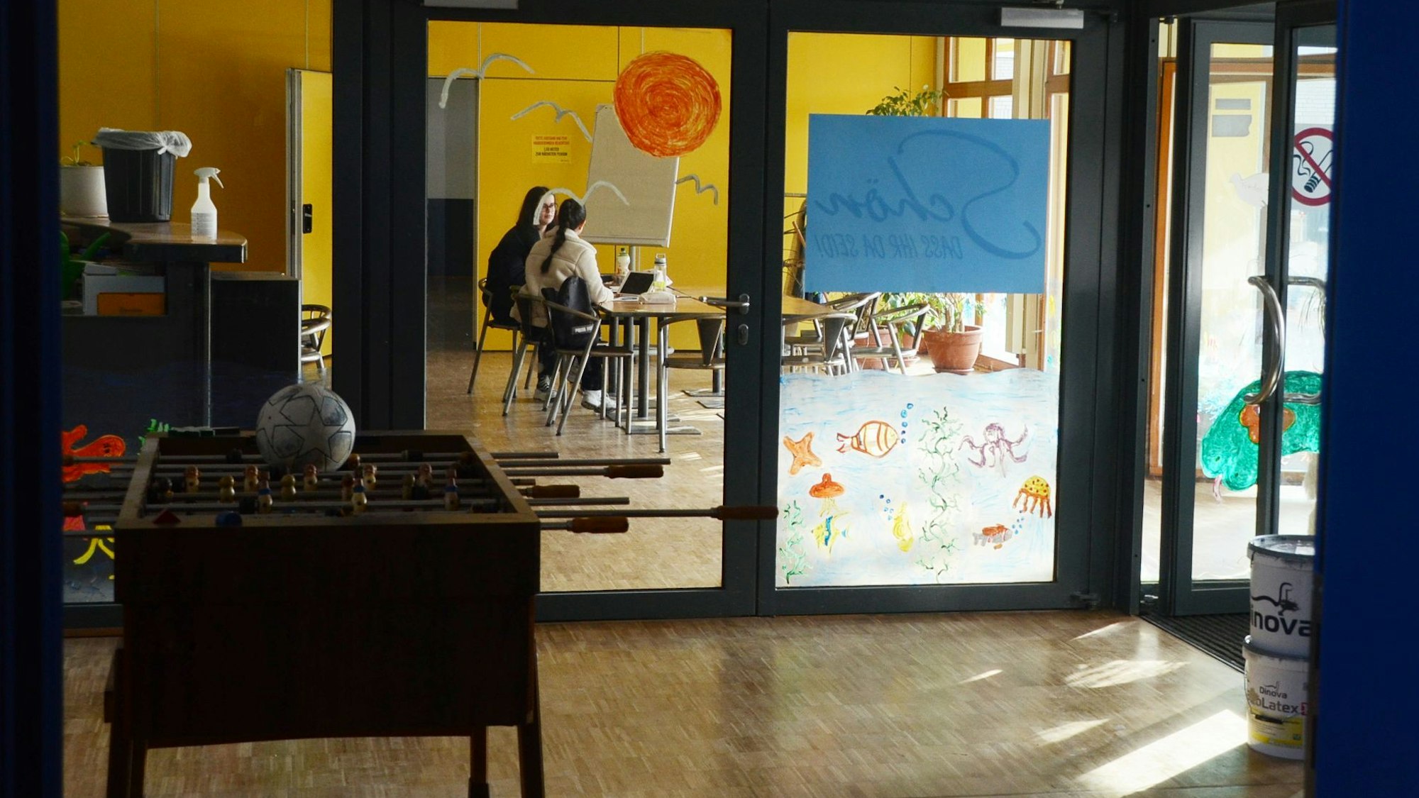 Ein Tischkicker vor einem Tischfußball, im Hintergrund sitzen zwei junge Frauen hinter einer Glastür.