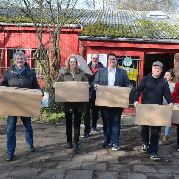 Männer und Frauen vor einem flachen roten Holzbau; sie tragen Umzugskisten.