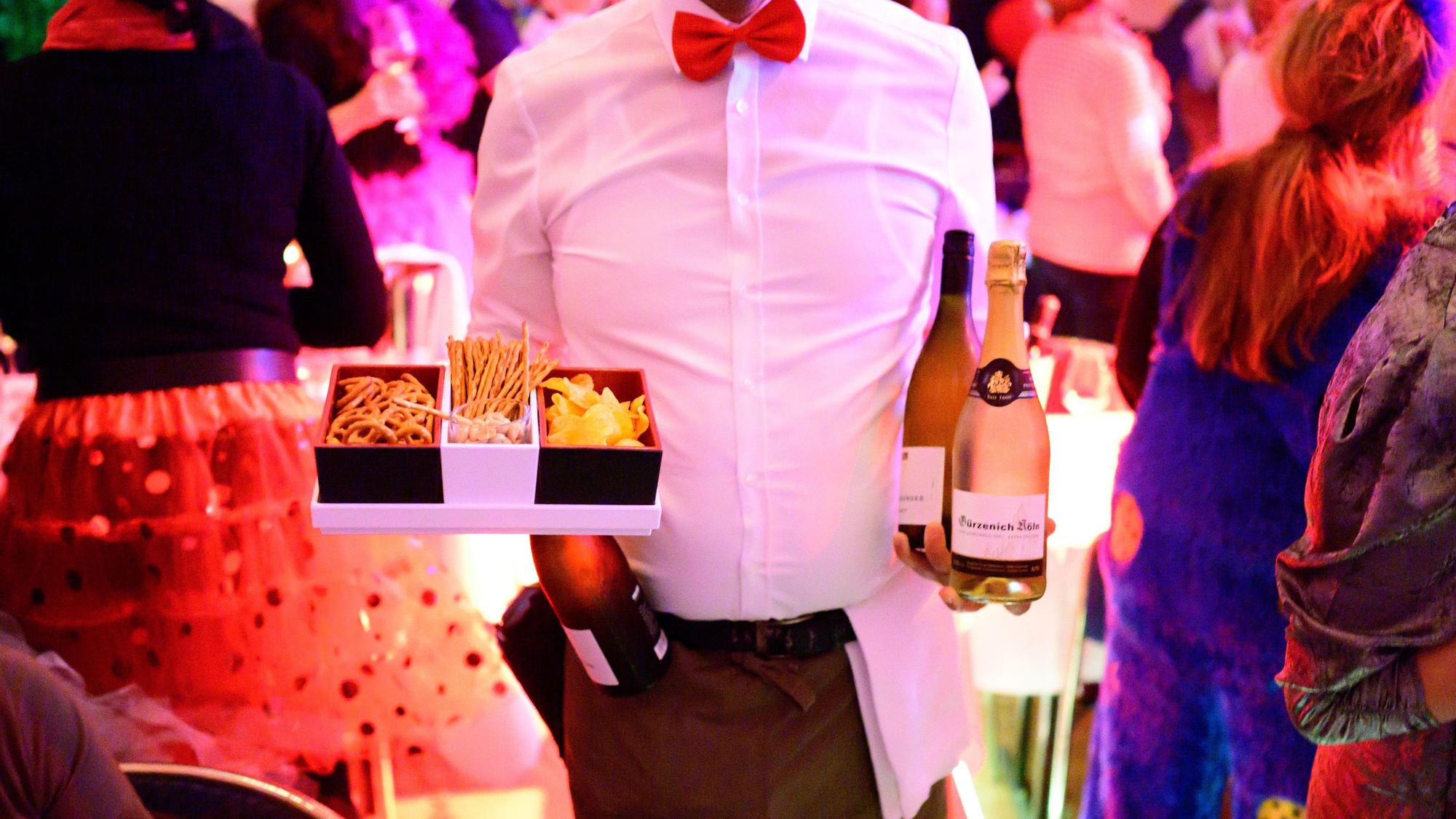 Ein Kellner hält eine Flasche Wein, eine Flasche Sekt und ein Tablett mit Chips, Salzstangen und Brezel in den Händen.