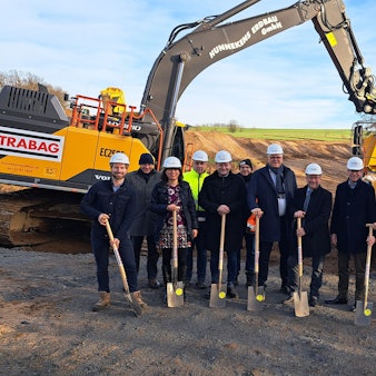 Das Foto zeigt eine Gruppe mit Bauarbeiterhelmen und Spaten, dahinter zwei Baufahrzeuge.