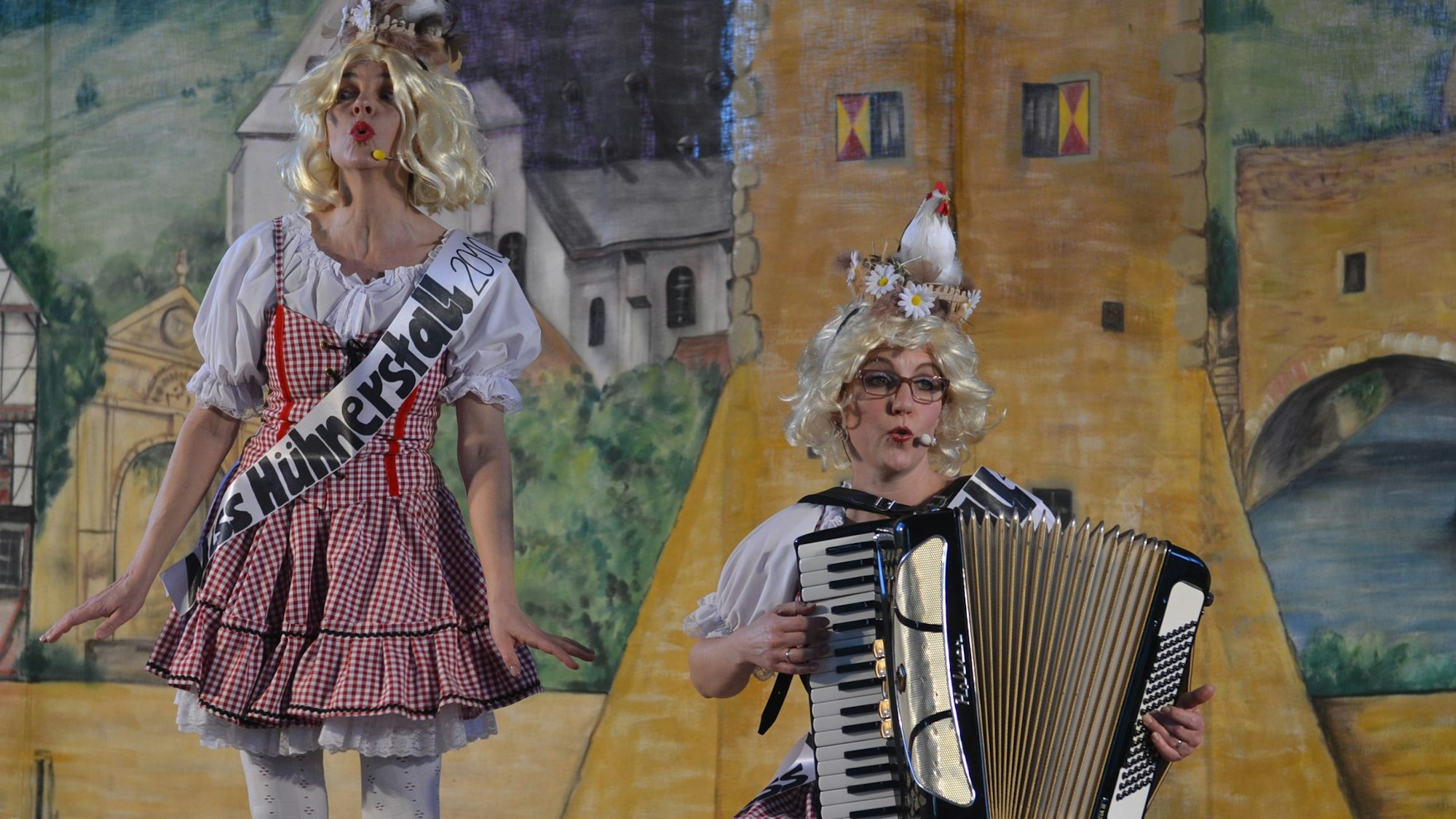 Zwei Frauen stehen im Dirndl und mit einem Huhn auf dem Kopf auf einer Bühne. Eine der beiden spielt Akkordeon, die andere trägt eine Schärpe auf der steht „Miss Hühnerstall 2010“