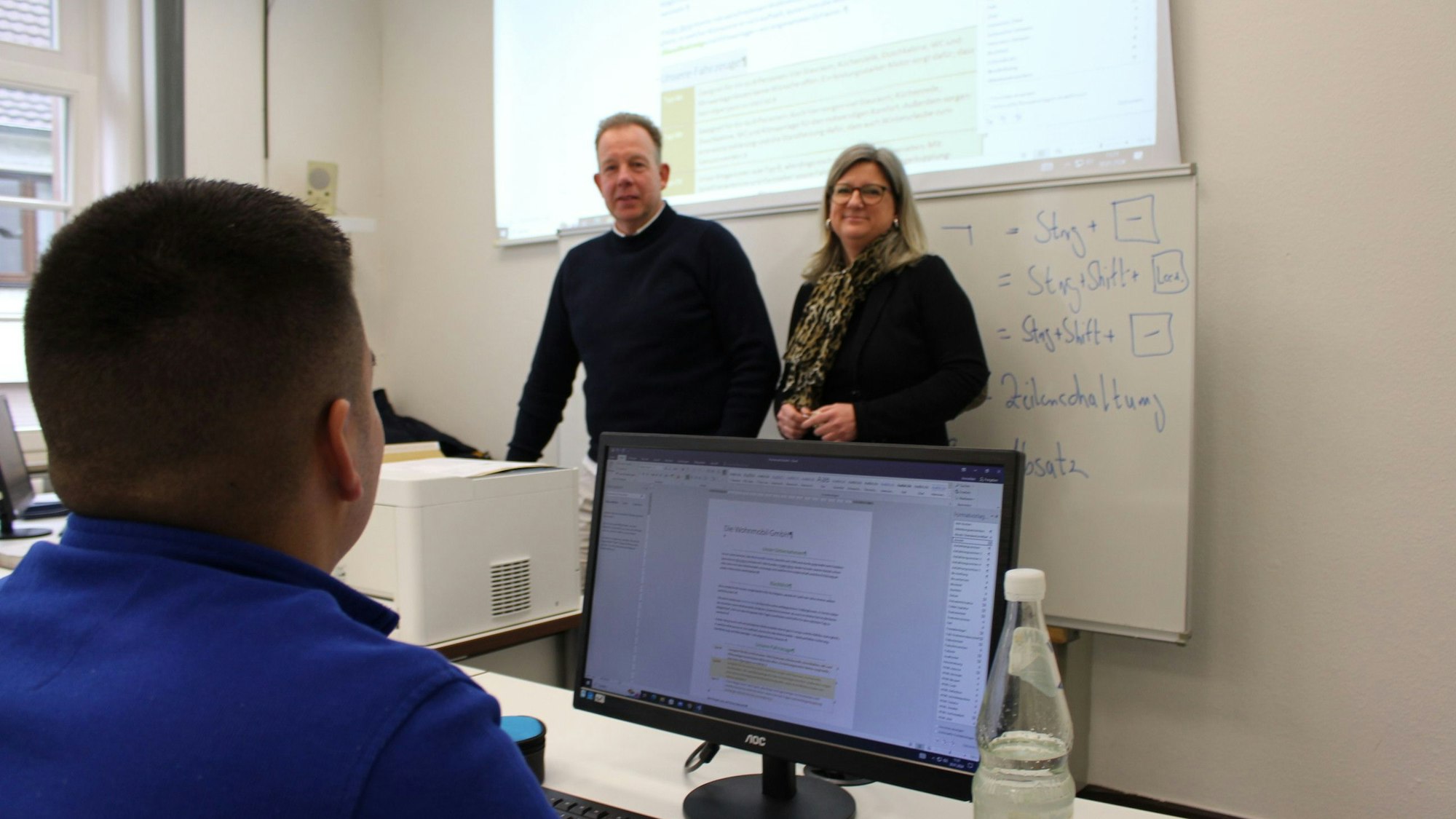 Vanessa Becker, Leiterin der Volkshochschule des Kreises Euskirchen, steht neben einem Kursleiter. Im Vordergrund sitzt ein Teilnehmer hinter einem Computerbildschirm.