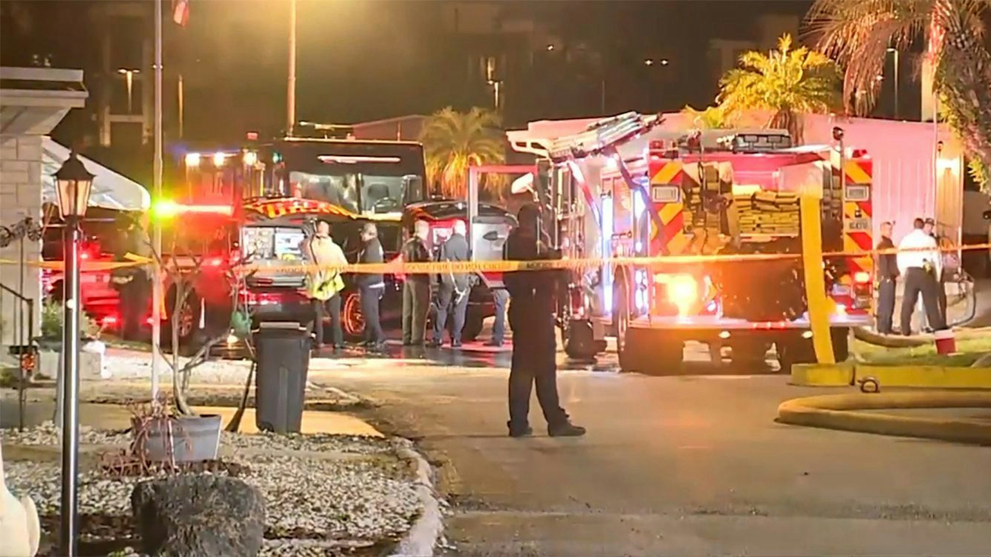 Mehrere Löschfahrzeuge mit Warnlichtern stehen bei Wohnungen und Palmen. (Screenshot)