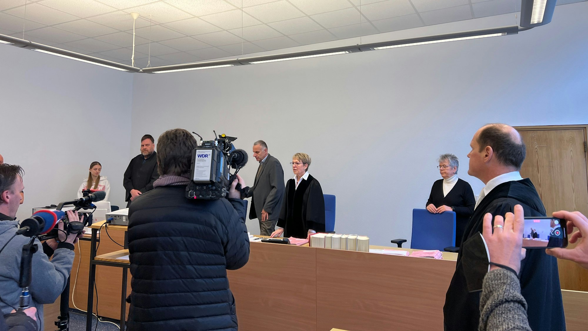 Journalisten filmen und fotografieren das Bensberger Schöffengericht vor Beginn des ersten Prozesses gegen einen der Kölner Nizza-Hooligans. In der Mitte  Schöffenrichterin Birgit Brandes, eingerahmt von Schöffe und Schöffin, rechts Oberstaatsanwalt Ulf Willuhn, der die Anklage vertrat.