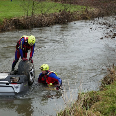 Zwei Männer in Neoprenanzügen stehen in einem Fluss an einem Auto, bei dem nur die Hinterräder aus dem Wasser ragen.