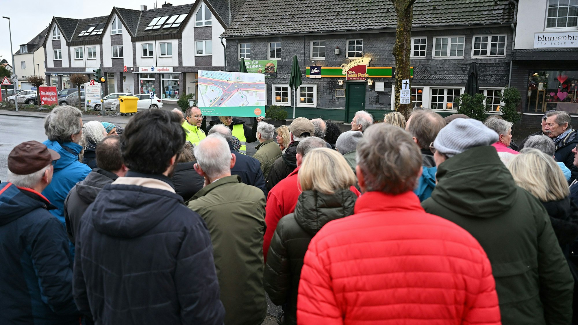 Stadtplaner halten ein Schild mit einer Zeichnung des geplanten Straßenverlaufs hoch. Drumherum stehen viele Menschen.