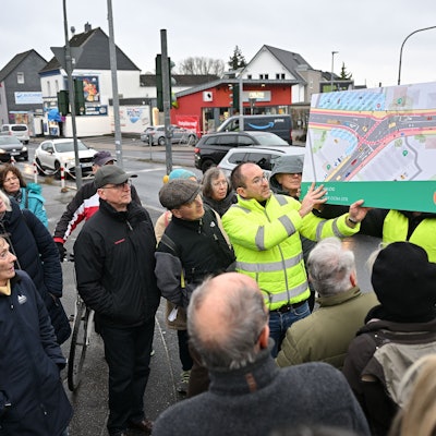 Ein Mitarbeiter des Planungsbüros in gelber Weste hält einen farbigen Entwurf, der die Gestaltung der Kreuzung zeigt, hoch. Teilnehmer der Veranstaltung stehen drumherum.