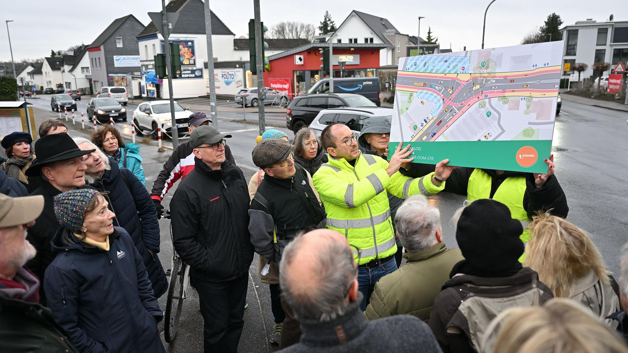 Ein Mitarbeiter des Planungsbüros in gelber Weste hält einen farbigen Entwurf, der die Gestaltung der Kreuzung zeigt, hoch. Teilnehmer der Veranstaltung stehen drumherum.