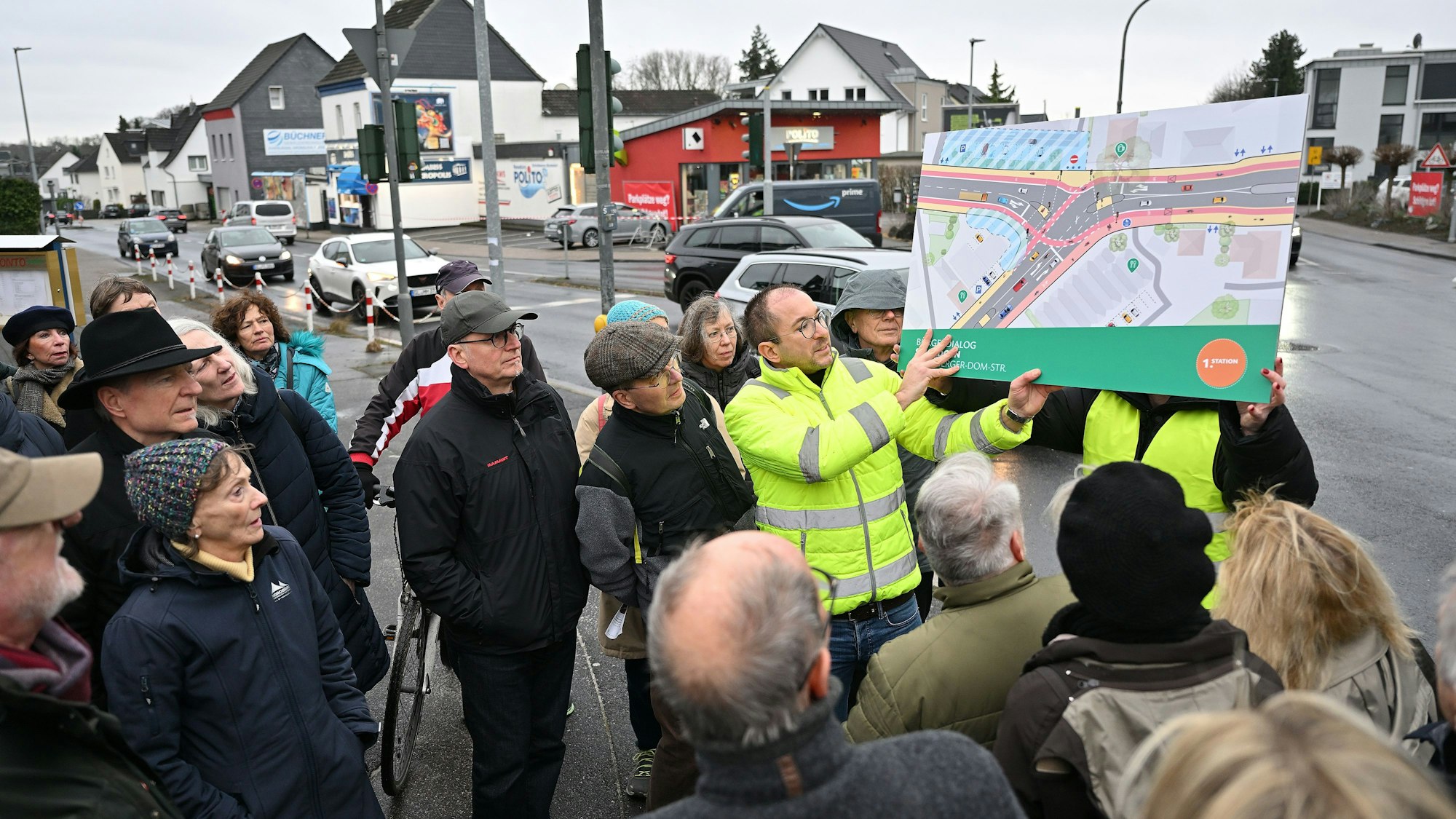 Ein Mitarbeiter des Planungsbüros in gelber Weste hält einen farbigen Entwurf, der die Gestaltung der Kreuzung zeigt, hoch. Teilnehmer der Veranstaltung stehen drumherum.