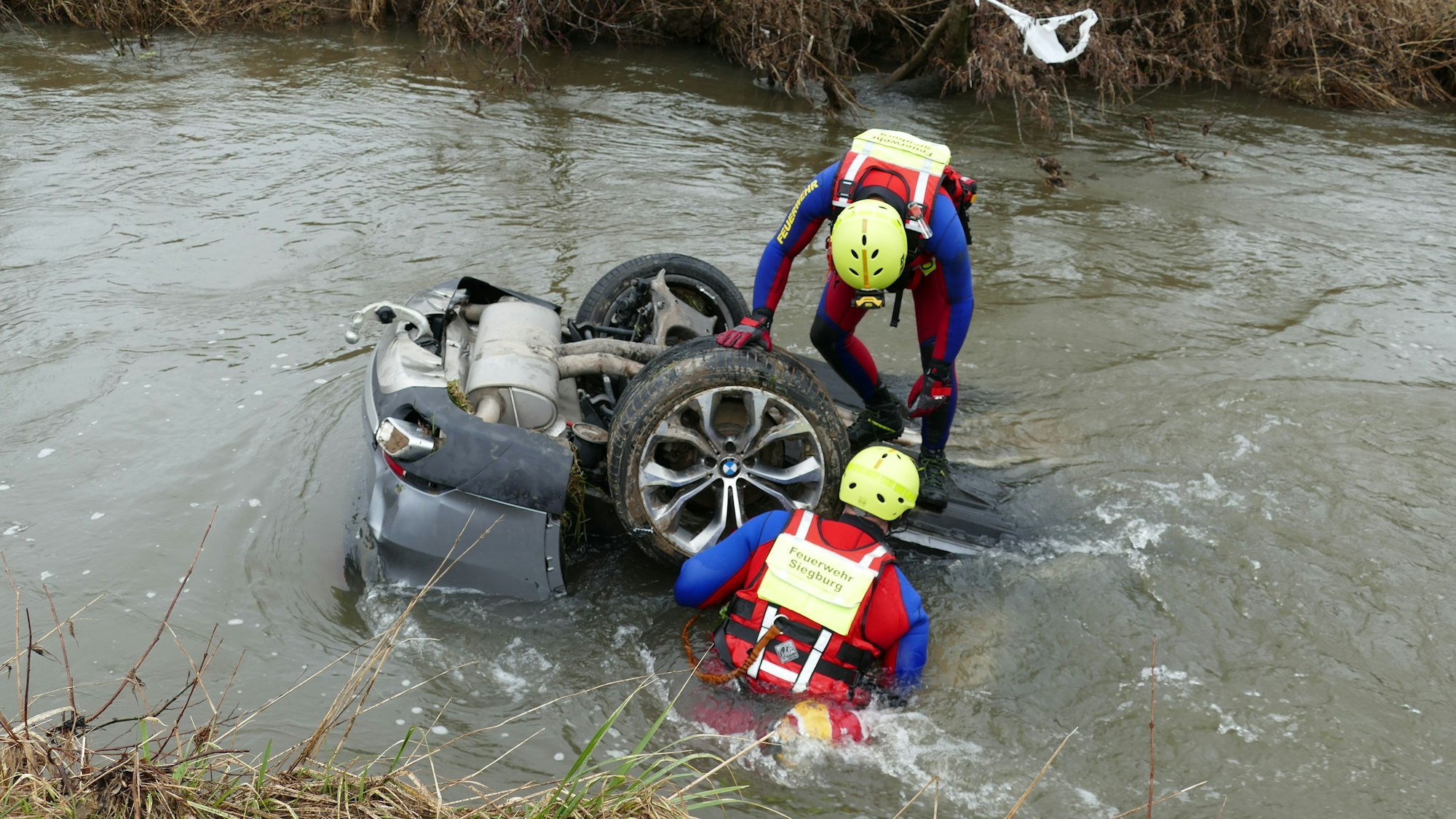 Zwei Feuerwehrleute stehen auf einem Auto, das auf dem Kopf im Wasser liegt.