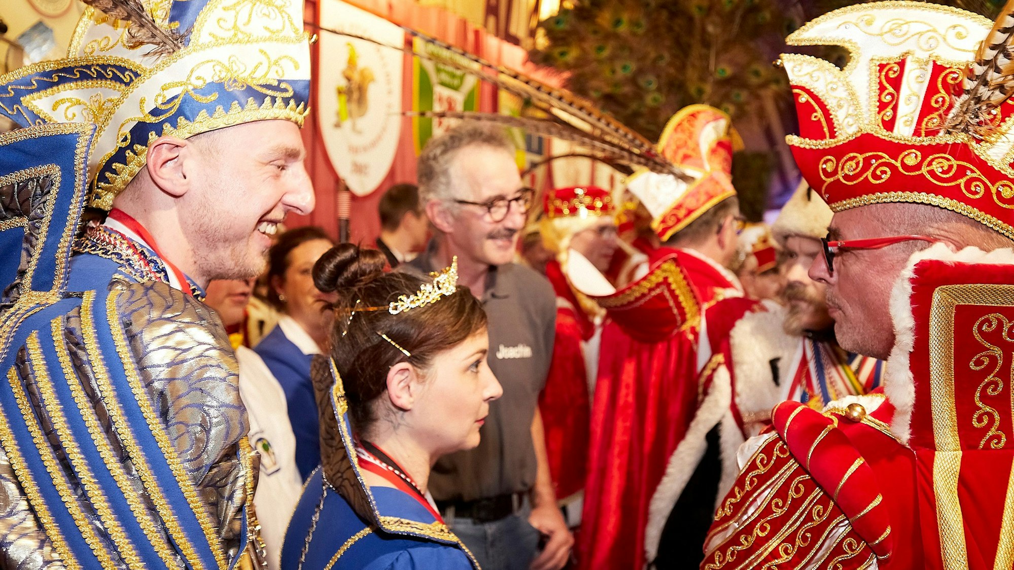 Zahlreiche Karnevals-Tollitäten stehen auf einer Bühne. Ein Prinz in rot-weißem Kostüm unterhält sich mit einem Prinzenpaar in blau-weißem Ornat.