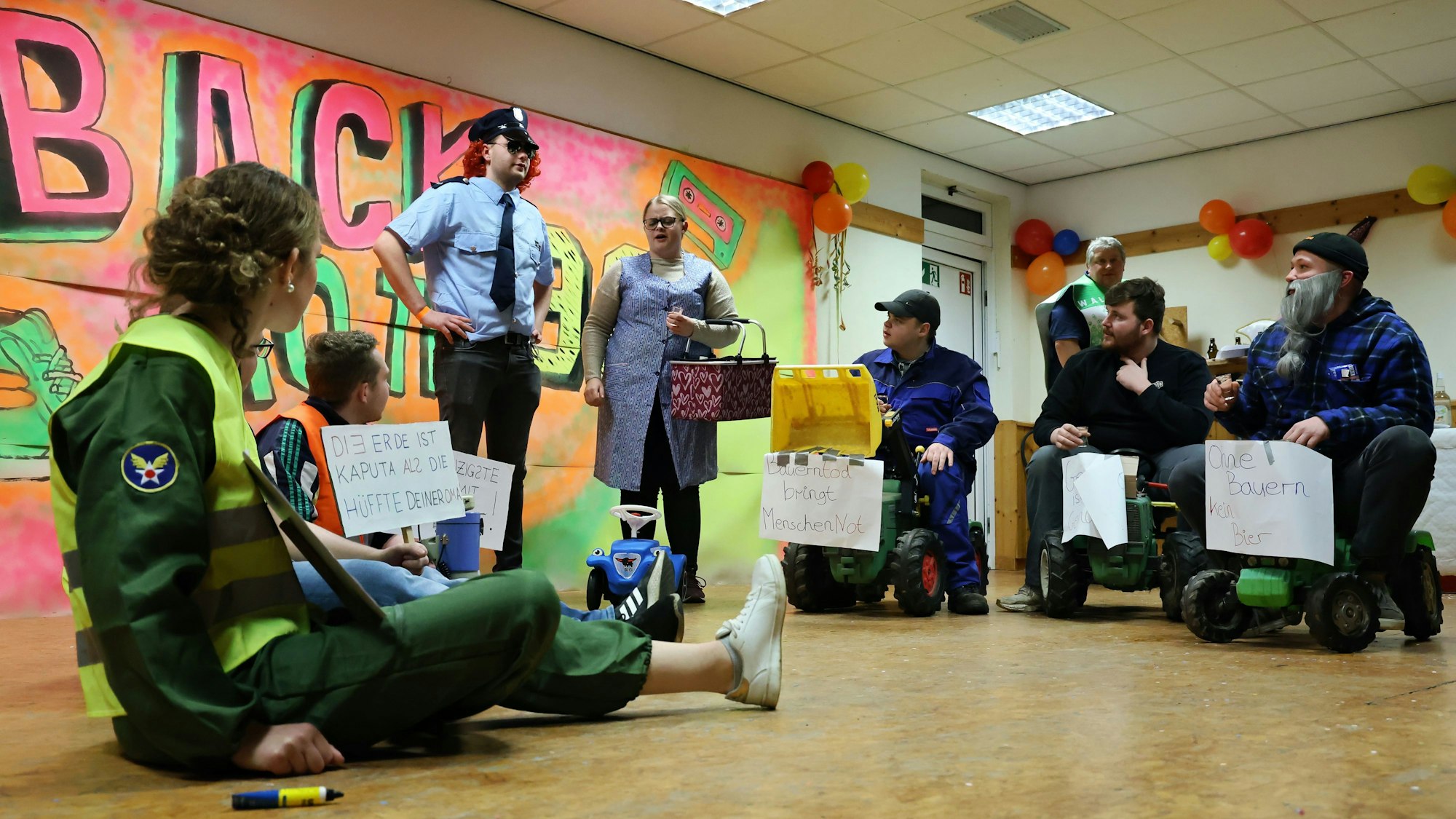Drei als Bauern verkleidete Akteure des Jugendclubs rollen auf drei sitzende Klimakleber zu. Alle haben Transparente. Ein Polizist und eine Bäuerin versuchen zu vermitteln.
