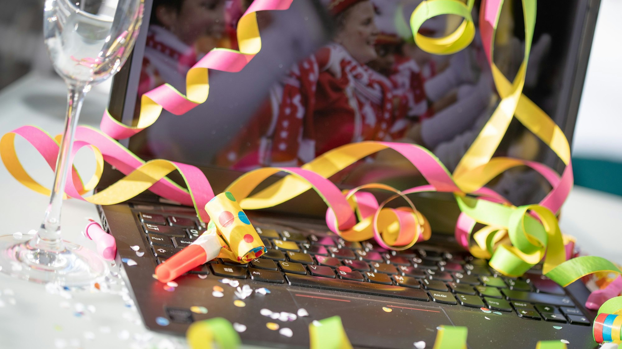 Das Foto einer Karnevalsgarde ist auf einem Notebook zu sehen. Davor liegen Konfetti, Luftschlangen, ein Glas und eine Tröte.