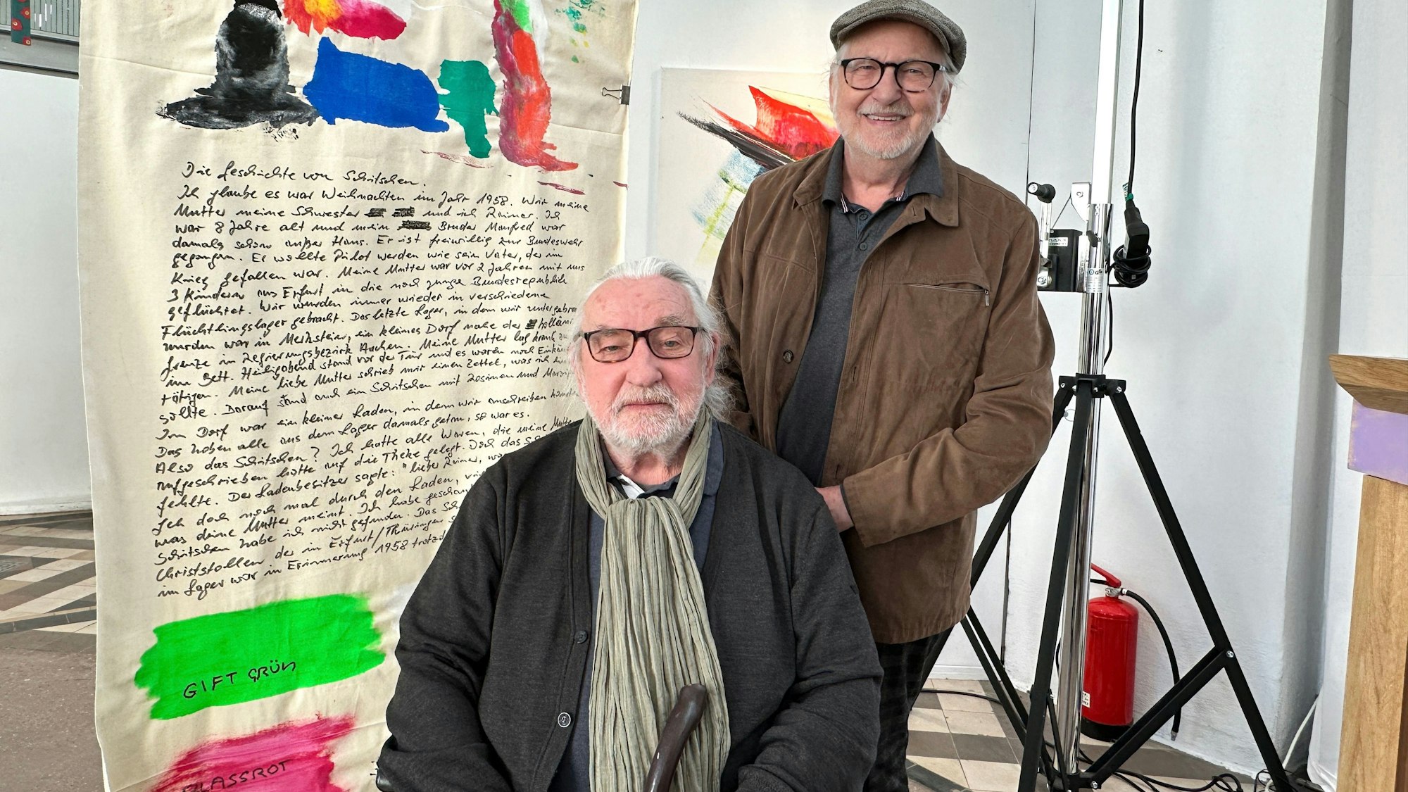 Zwei Männer (einer davon im Rollstuhl) stehen neben einer bemalten und beschriebenen Stoffbahn.