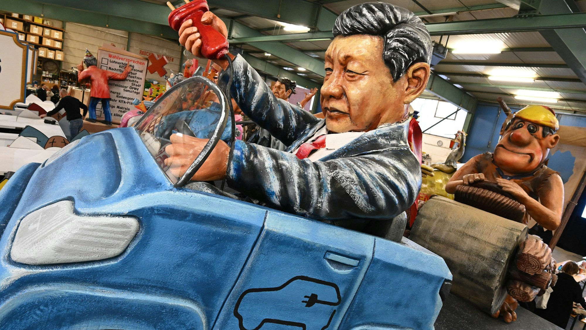 06.02.2024, Rheinland-Pfalz, Mainz: Chinas Staats- und Parteichef Xi Jinping im Elektroauto und der deutsche Michel als ·Steinzeit-Fred Feuerstein· werden bei der Vorstellung der Motivwagen für den Mainzer Rosenmontagszug in der Wagenhalle des Mainzer Carneval-Vereins (MCV) präsentiert. Innen- und außenpolitischen Themen dienen als Vorlage für die närrischen Motivwagen, die am 12. Februar im närrischen Lindwurm durch die Landeshauptstadt rollen. Foto: Arne Dedert/dpa +++ dpa-Bildfunk +++