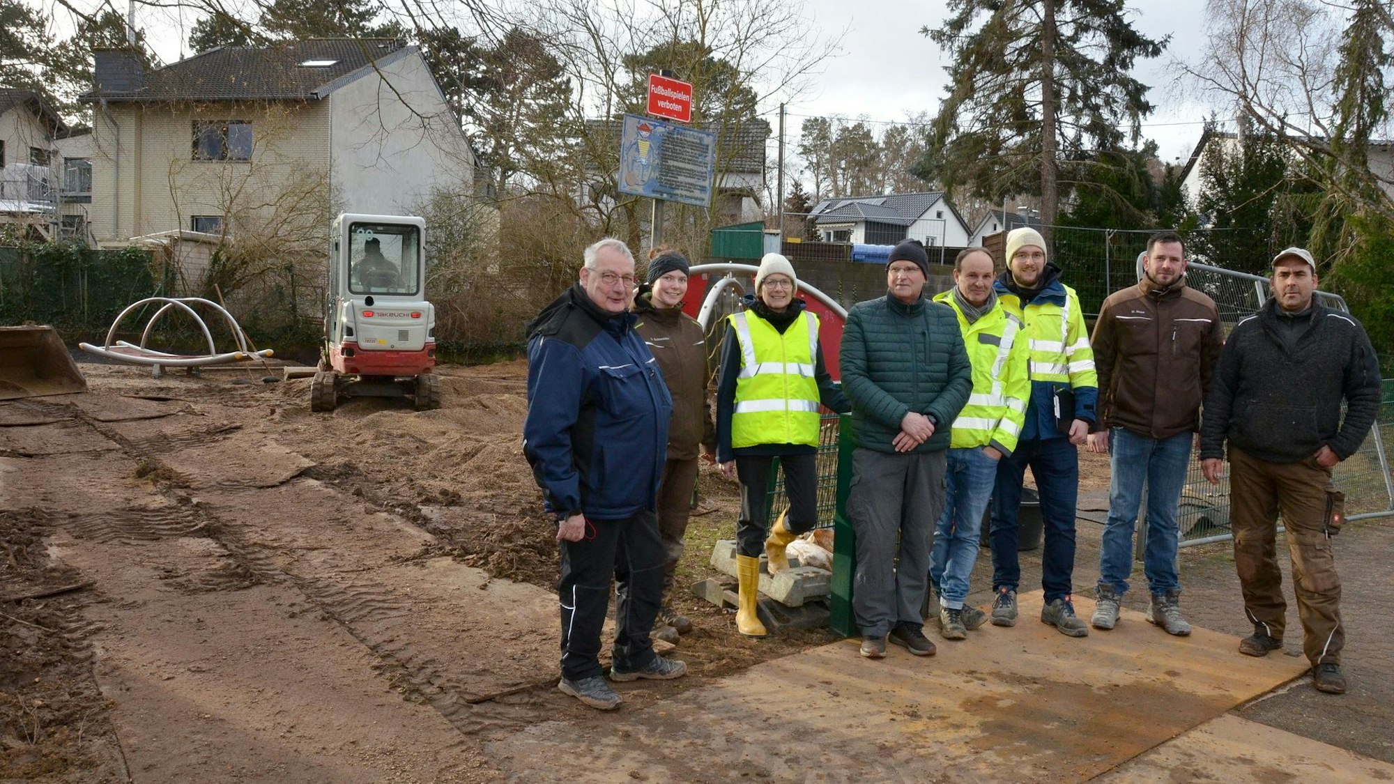 Während ein Mini-Bagger auf dem Spielplatzgelände arbeitet, haben sich Vertreter von Behörden und Firmen zum Gruppenbild aufgestellt.