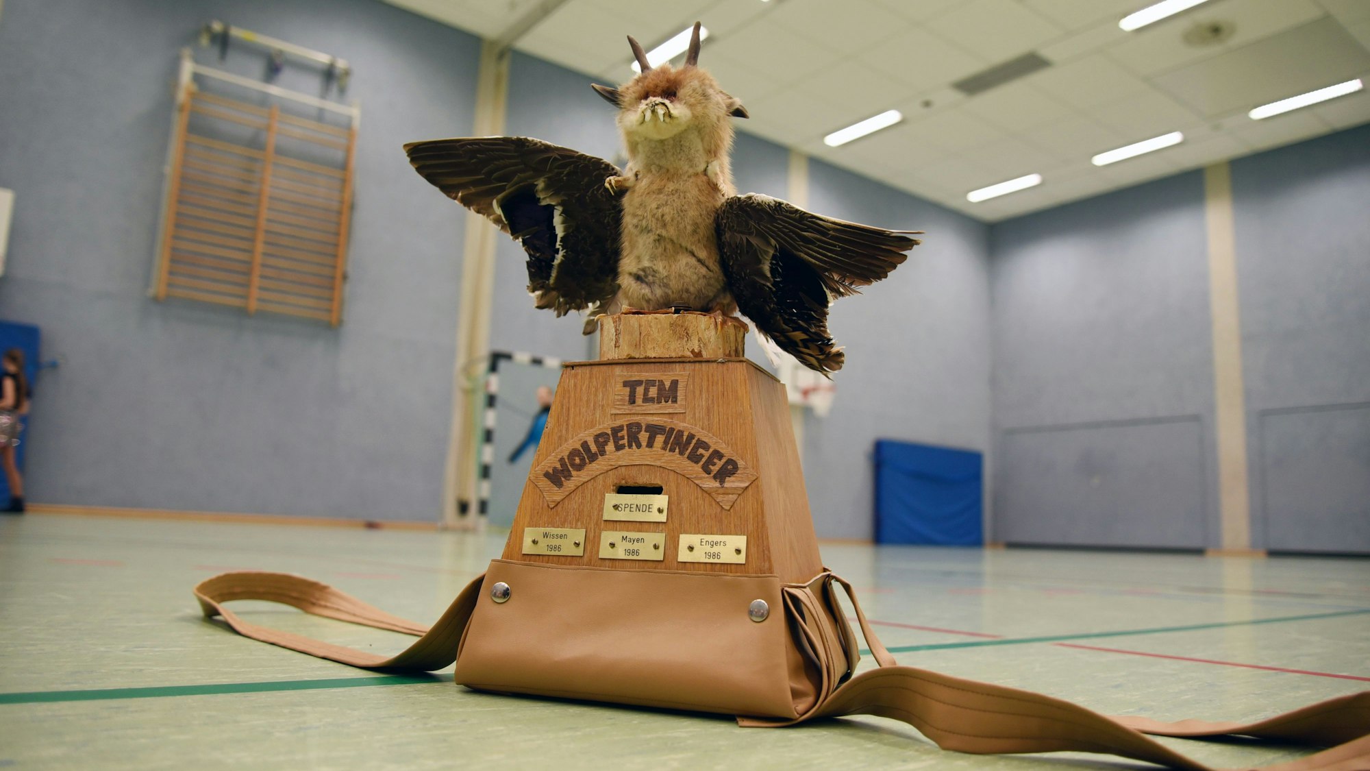 Ein tierisches Fabelwesen mit Flügeln steht auf einem Holzpodest. Auf diesem steht: „Die Wolpertinger“.
