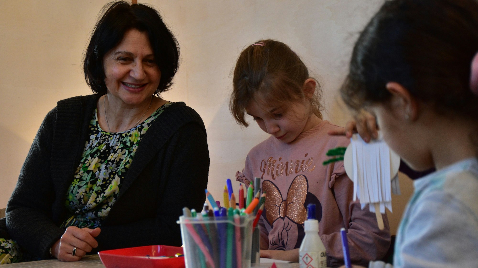 Zwei Mädchen basteln; eine erwachsene Betreuerin schaut zu.