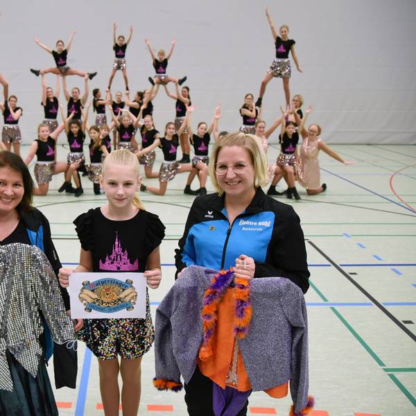Zwei Frauen und ein Mädchen stehen zusammen. Die beiden Frauen halten Tanzkostüme in den Händen. Im Hintergrund ist eine Tanzgruppe zu sehen.