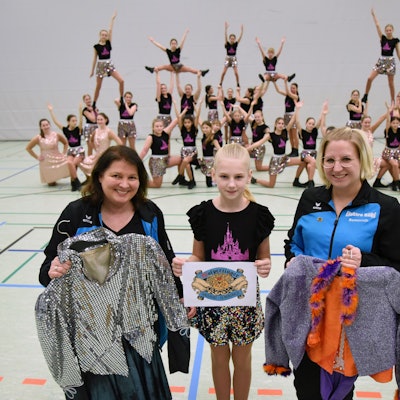 Zwei Frauen und ein Mädchen stehen zusammen. Die beiden Frauen halten Tanzkostüme in den Händen. Im Hintergrund ist eine Tanzgruppe zu sehen.