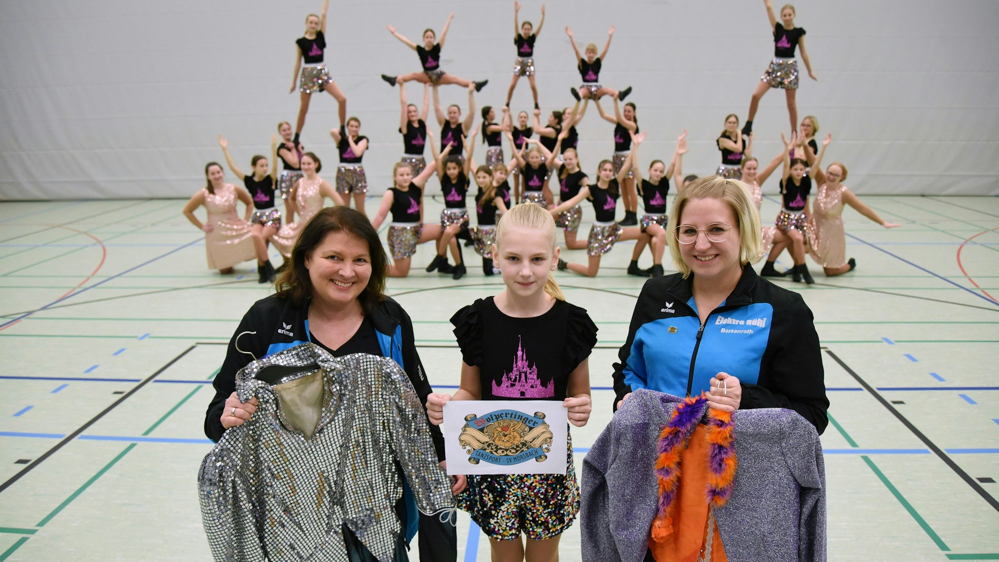 Zwei Frauen und ein Mädchen stehen zusammen. Die beiden Frauen halten Tanzkostüme in den Händen. Im Hintergrund ist eine Tanzgruppe zu sehen.