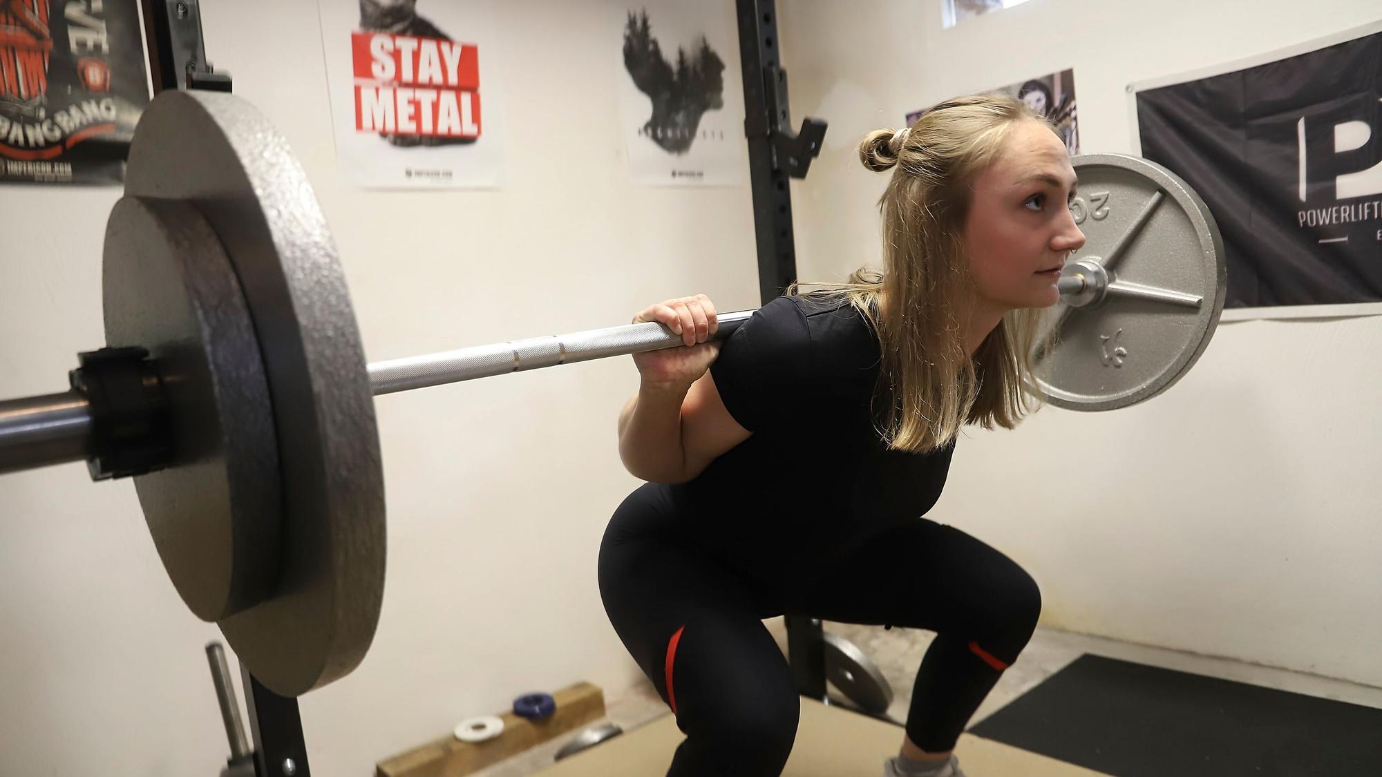 Beim Training stemmt Helena Decker Gewichte. Dabei geht sie in die Knie. Auf einem Poster im Hintergrund steht „Stay Metal“.