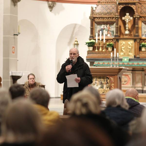 Ein Geistlicher in schwarzem Anorak steht in einer voll besetzten Kirche und spricht in ein Mikrofon, eine Frau sitzt daneben.