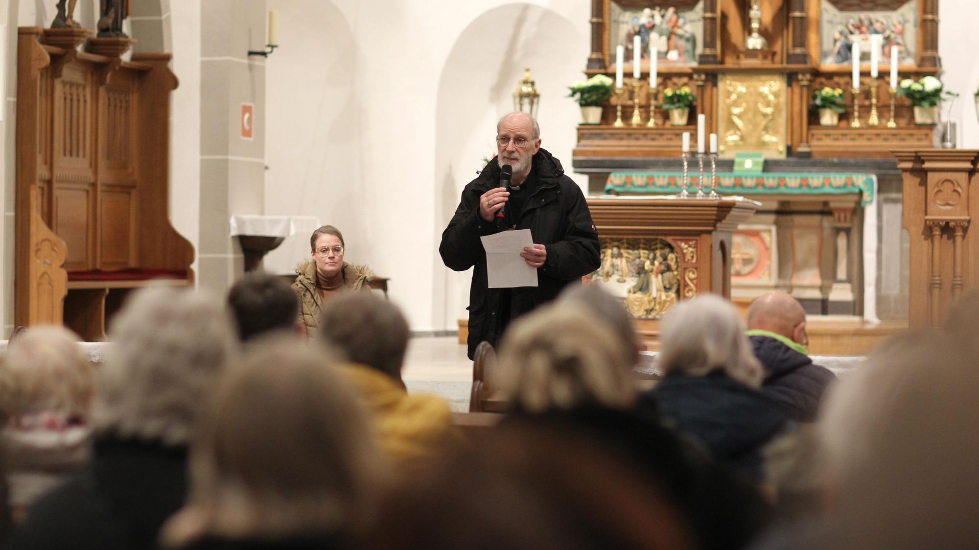 Ein älterer Geistlicher mit Mikrofon und einem Papier in der Hand steht in einer voll besetzten Kirche.