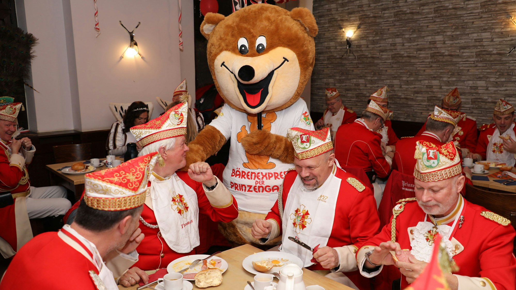 Ein Mensch im Bärenkostüm steht zwischen Prinzengardisten, die frühstücken.