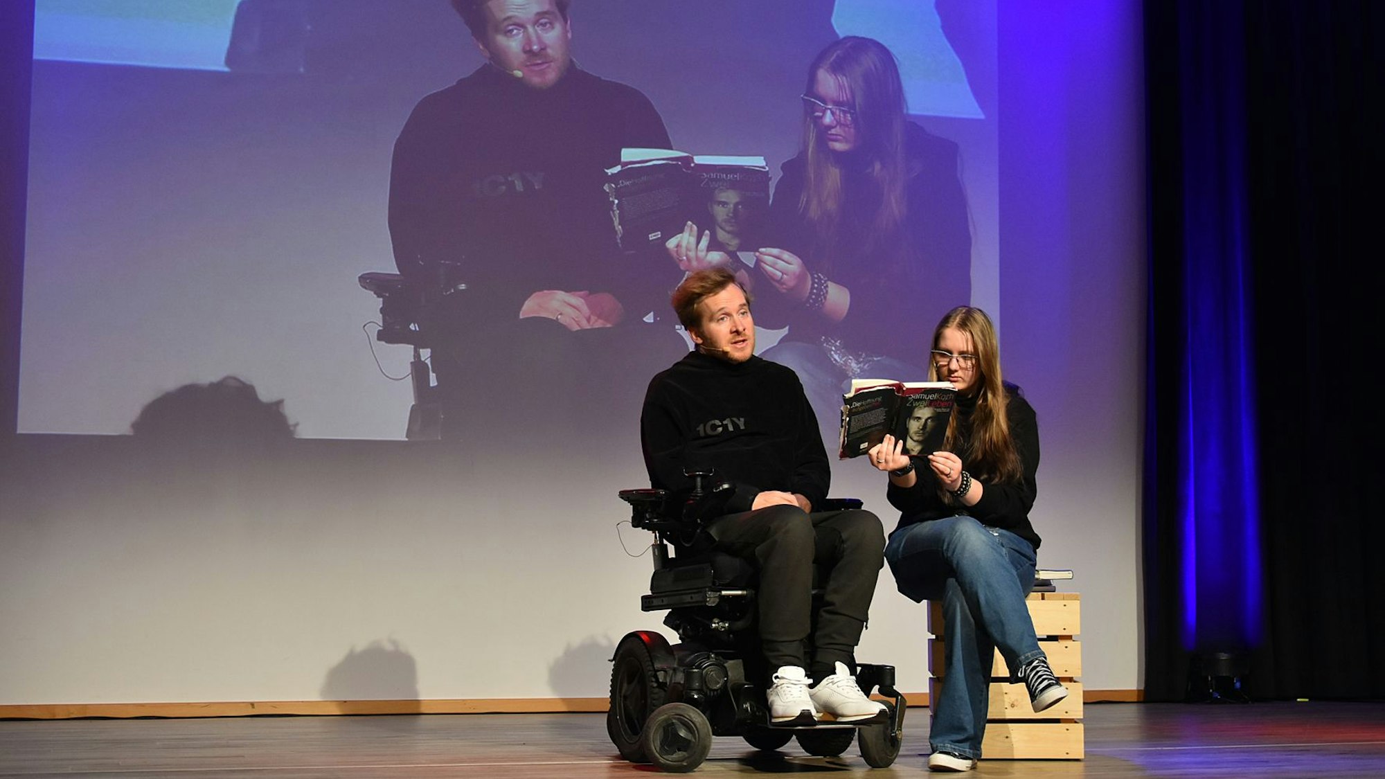 Samuel Koch (im Rollstuhl) auf der Bühne mit einer Schülerin, die ihm ein Buch hinhält.