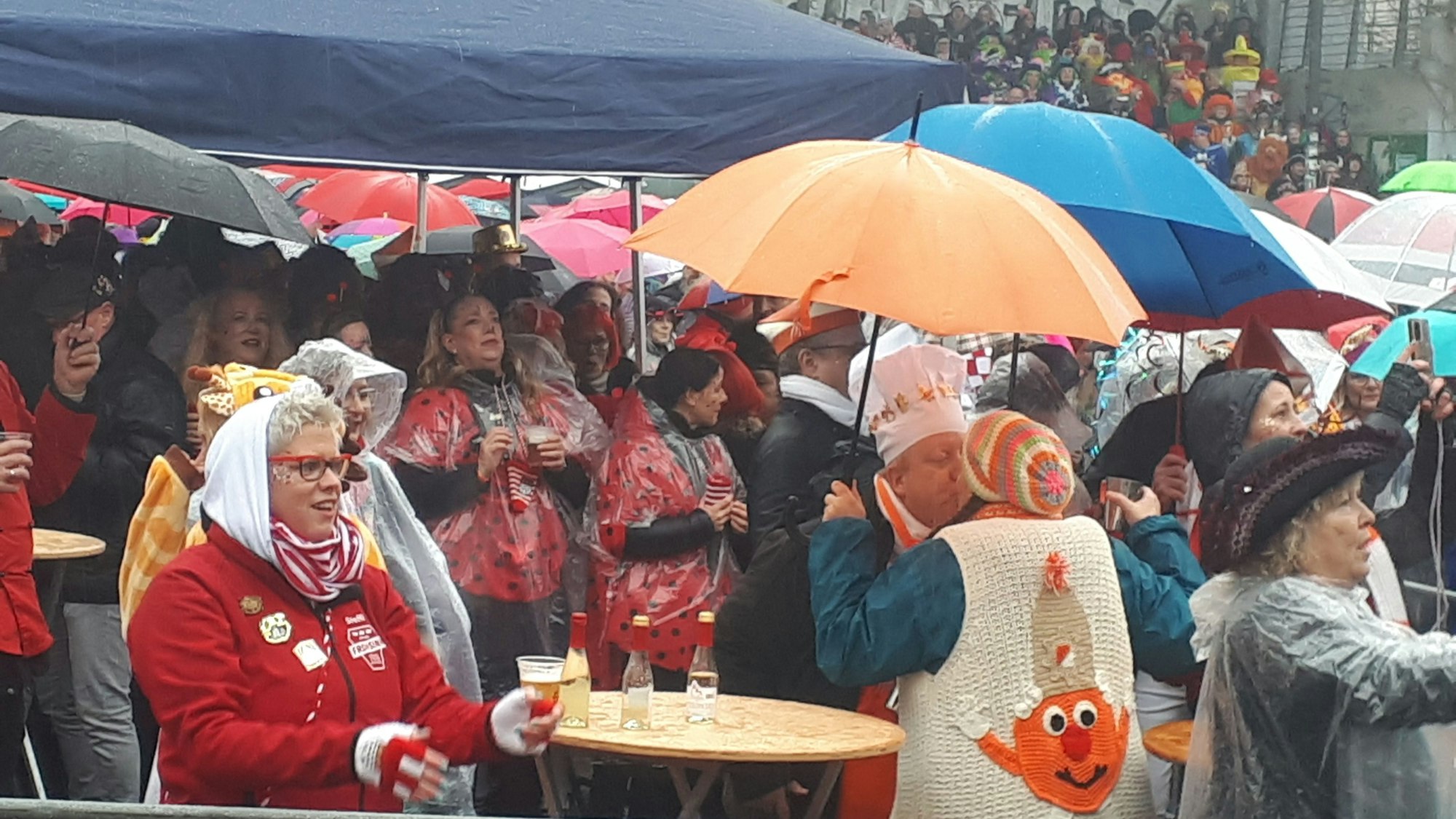 Menschen mit Schirmen stehen auf einem Platz. Auch ein Pavillon als Regenschutz ist aufgebaut.