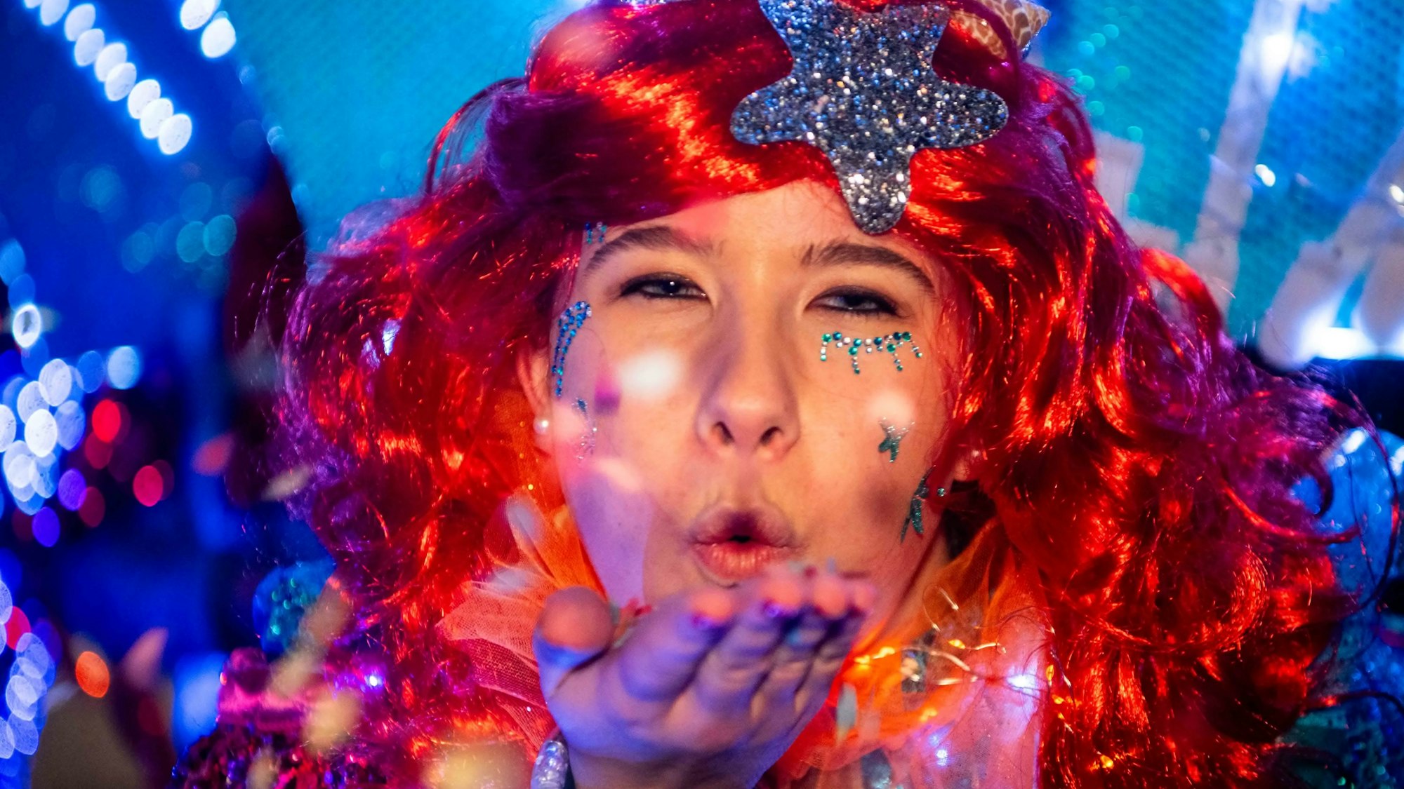 Eine Karnevalistin mit einer roten Perücke und einem silbernen Stern im Haar wirft den Jecken beim Lichterzug in Eiserfey eine Kusshand zu.