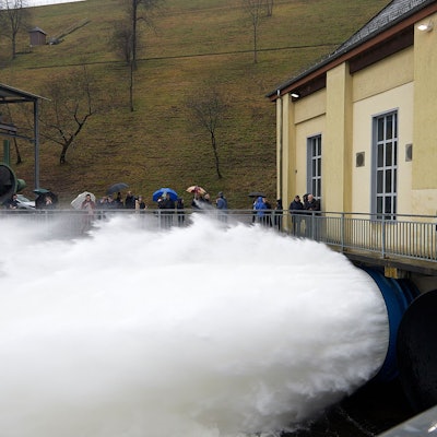 Aus großen Rohren unter einem gelben Gebäude schießt Wasser. Drumherum stehen Menschen mit Regenschirmen.