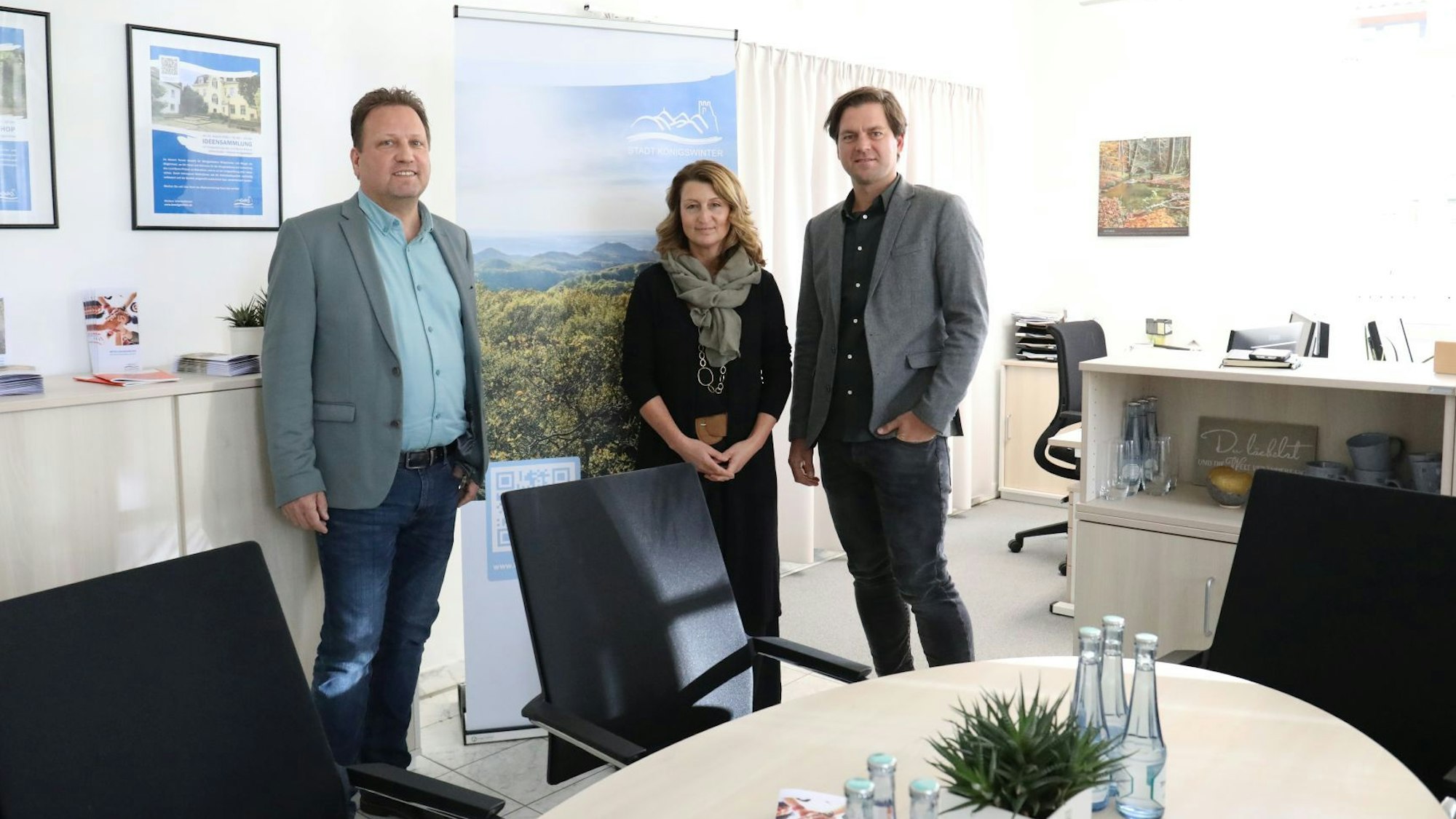 Zwei Männer und eine Frau stehen in einem Büro vor einem Tisch.