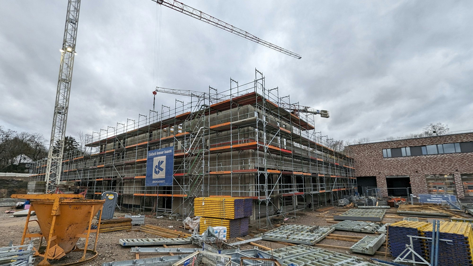 Ein eingerüsteter Rohbau mit Baumaterial und Baukran vor dem bereits fertiggestellten Gebäudetrakt der neuen Feuer- und Rettungswache.