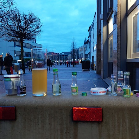Leere Schnapsfläschchen und ein halbleeres Bierglas, bunte Flaschendeckel auf einer Betonsperre.