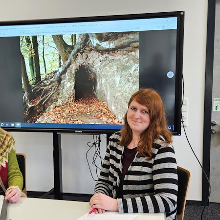Katrin Oberländer (l.) und Bibliotheksleiterin Michelle Wagner sitzen an einem Tisch. Im Hintergrund ist ein Foto vom Römerkanal zu sehen.