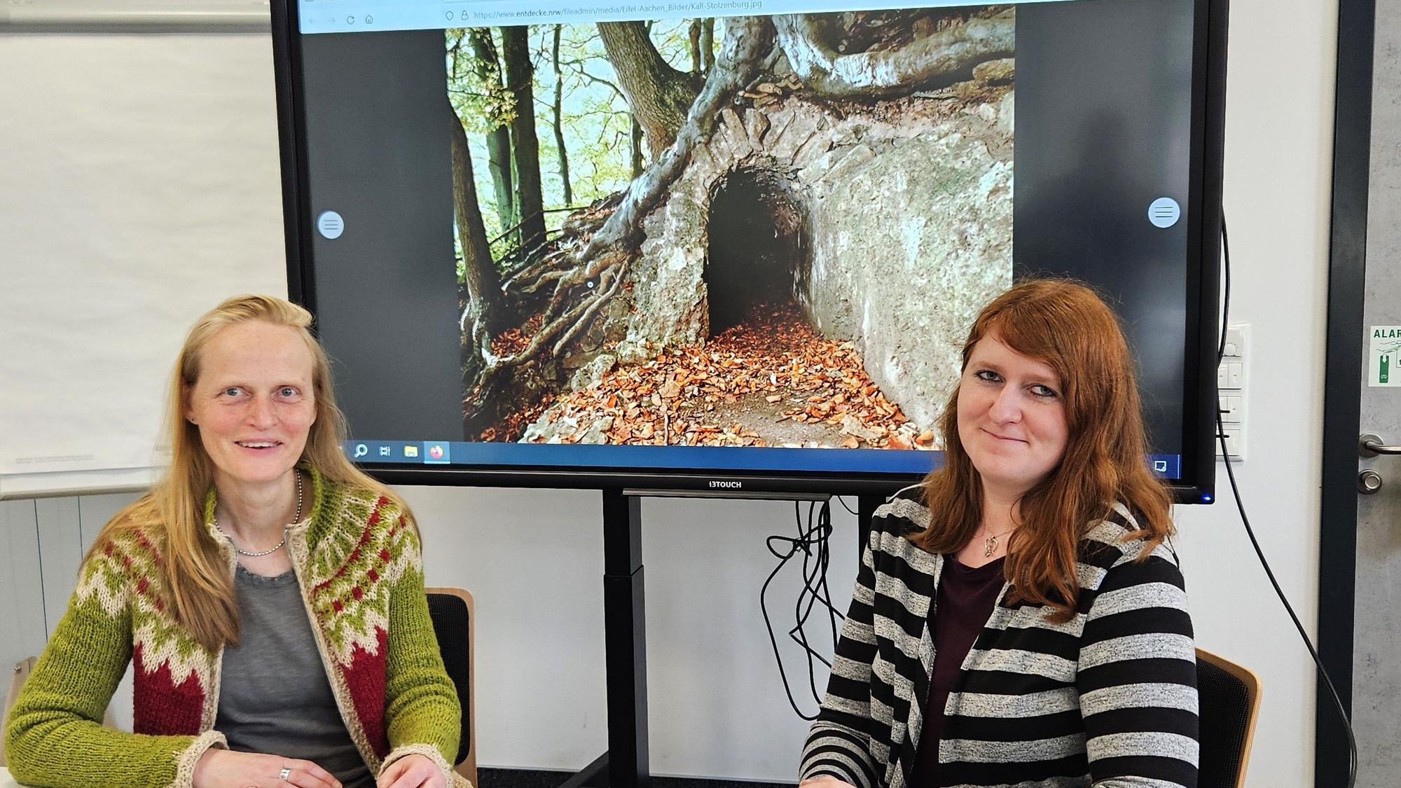 Katrin Oberländer (l.) und Bibliotheksleiterin Michelle Wagner sitzen an einem Tisch. Im Hintergrund ist ein Foto vom Römerkanal zu sehen.