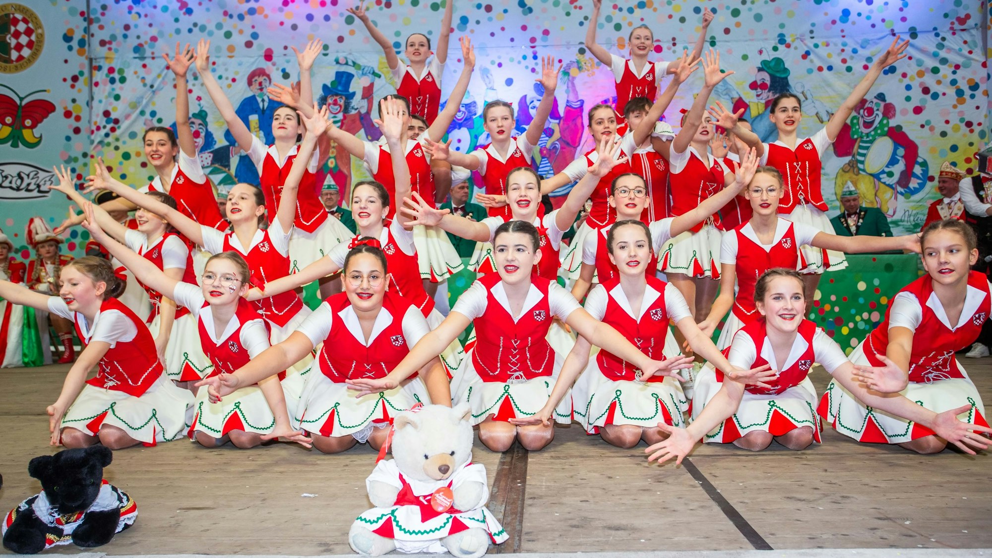 Eine Tanzgruppe auf der Bühne im Rahmen einer Karnevalssitzung in einem Festzelt. Vor ihnen sitzt ein weißer Plüschteddy am Rand der Bühne.