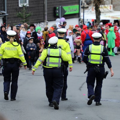 Polizisten gehen auf der Kürtener Bergstraße, auf der der Karnevalszug herannaht. Am Rand stehen kostümierte Jecke.