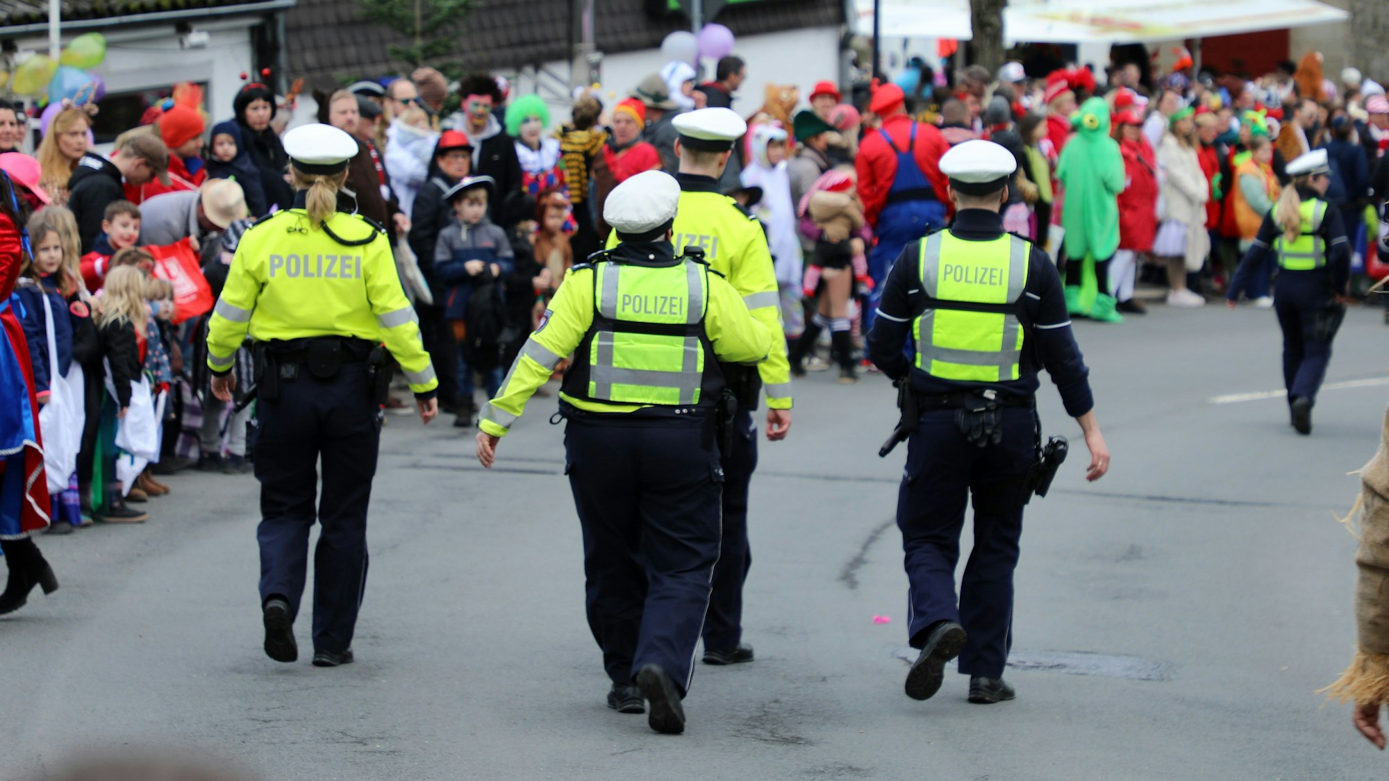 Polizisten gehen auf der Kürtener Bergstraße, auf der der Karnevalszug herannaht. Am Rand stehen kostümierte Jecke.