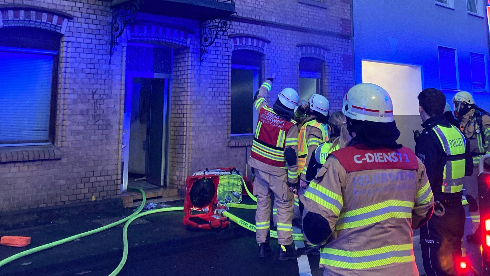 Feuerwehrleute stehen vor einem Haus, in das ein Feuerwehrschlauch führt. Vor der Eingangstür steht ein Lüfter.