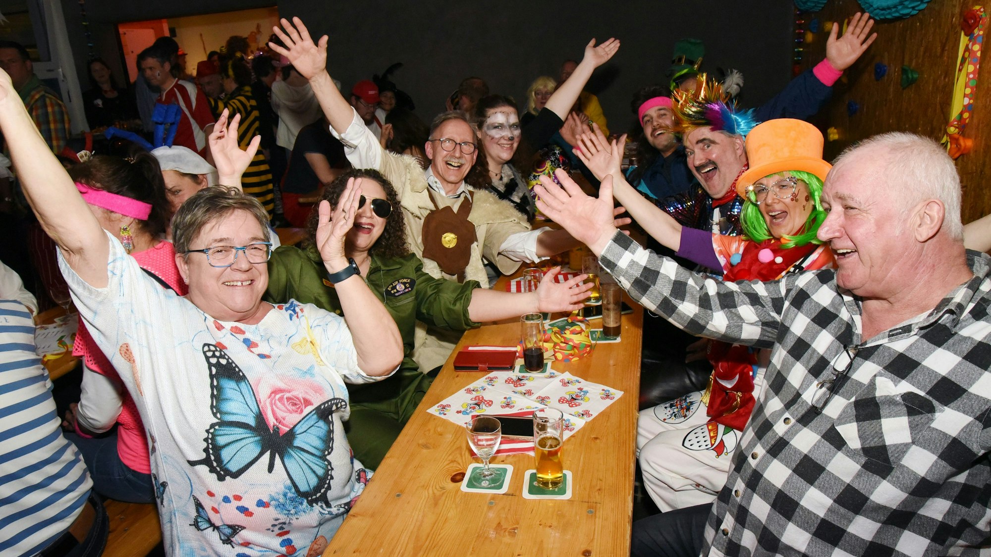 Mehrere verkleidete Leute sitzen gemeinsamen an einem Tisch, schauen in die Kamera und reißen jubelnd die Hände in die Luft.