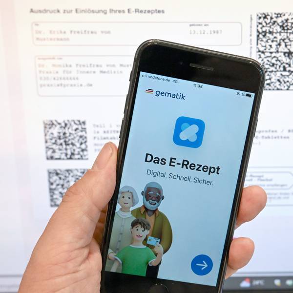Eine App, die das E-Rezept anzeigen kann, wird in einer Apotheke vor einem Bildschirm gehalten, der Hinweise zu den E-Rezepten anzeigt.