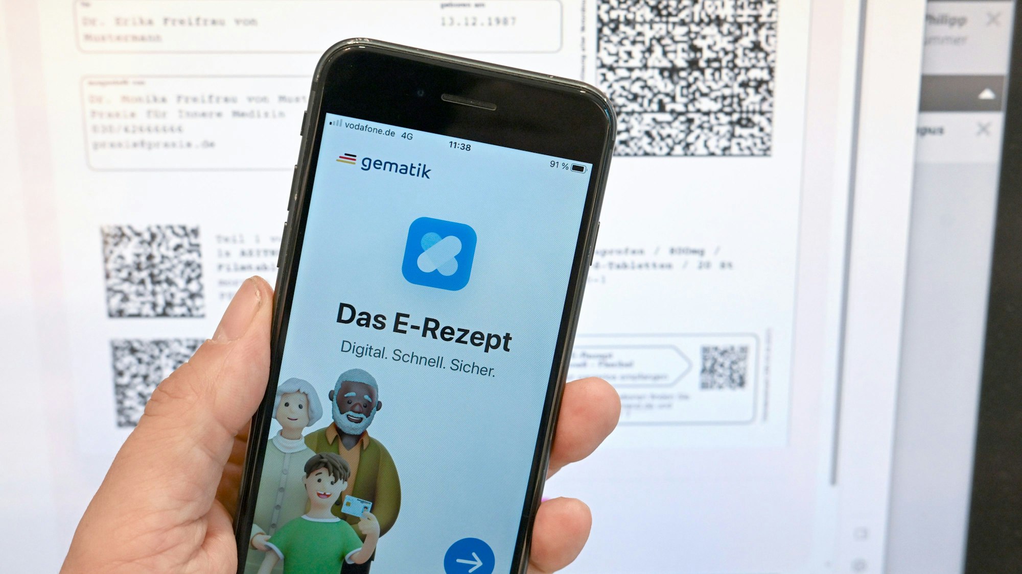 Eine App, die das E-Rezept anzeigen kann, wird in einer Apotheke vor einem Bildschirm gehalten, der Hinweise zu den E-Rezepten anzeigt.