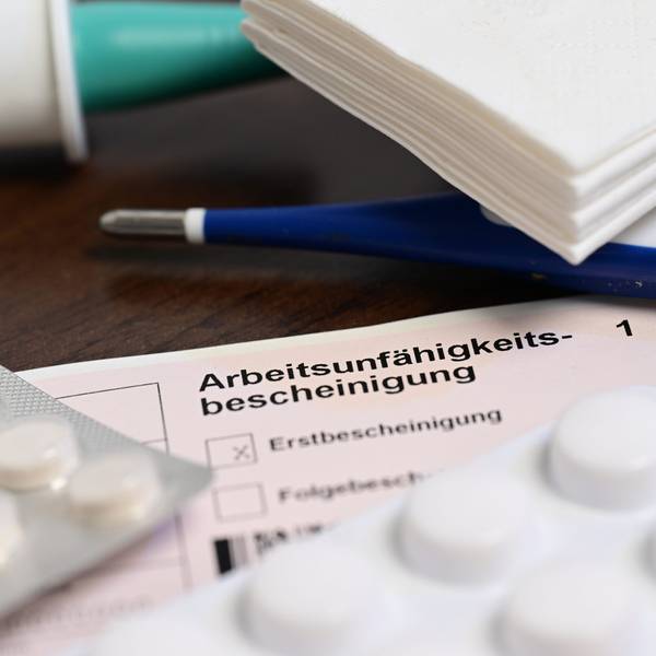 Eine Arbeitsunfähigkeitsbescheinigung liegt zwischen Taschentüchern, Tabletten und einem Fieberthermometer auf einem Tisch